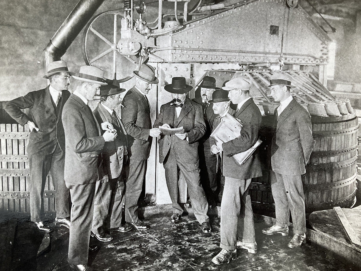 roupe d'hommes en costumes et chapeaux des années 1900 discutant près d'un pressoir.