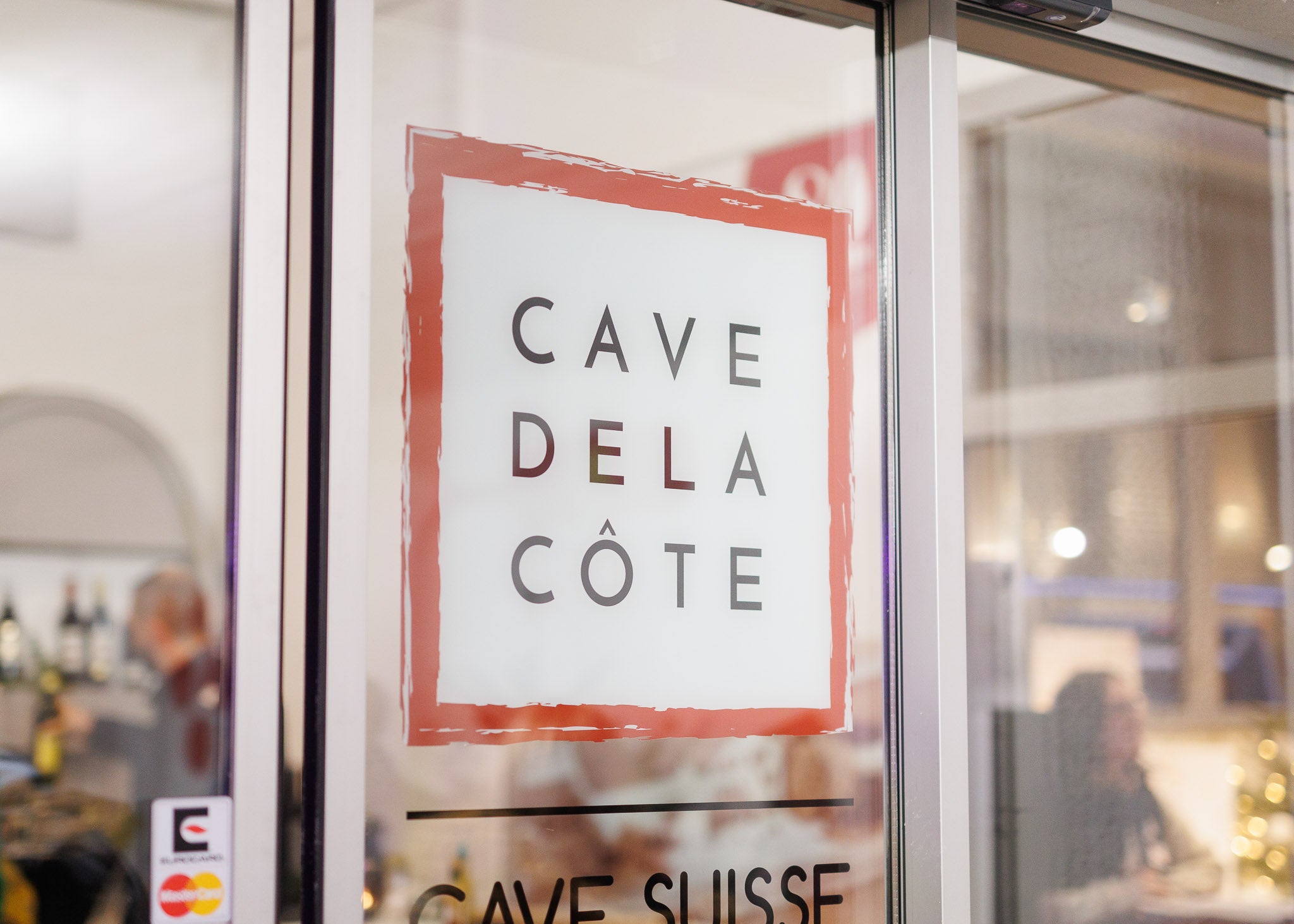 Vitrine de la Cave de La Côte avec un logo rouge et blanc 'CAVE DE LA CÔTE' collé sur la porte. À travers la vitre, on aperçoit l'intérieur du magasin, où des bouteilles de vin et des étagères sont visibles.