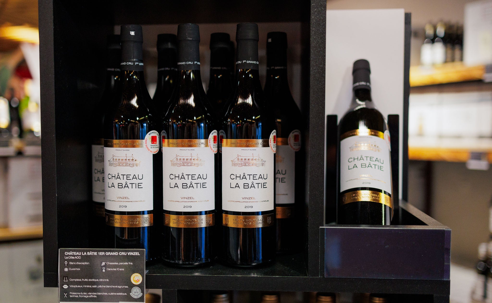 Présentoir en magasin mettant en avant plusieurs bouteilles de vin 'Château La Bâtie' (appellation Vinzel, 1er Grand Cru). Les bouteilles sont alignées avec des étiquettes blanches et dorées. Une fiche descriptive est visible en bas à gauche, indiquant les détails du vin.