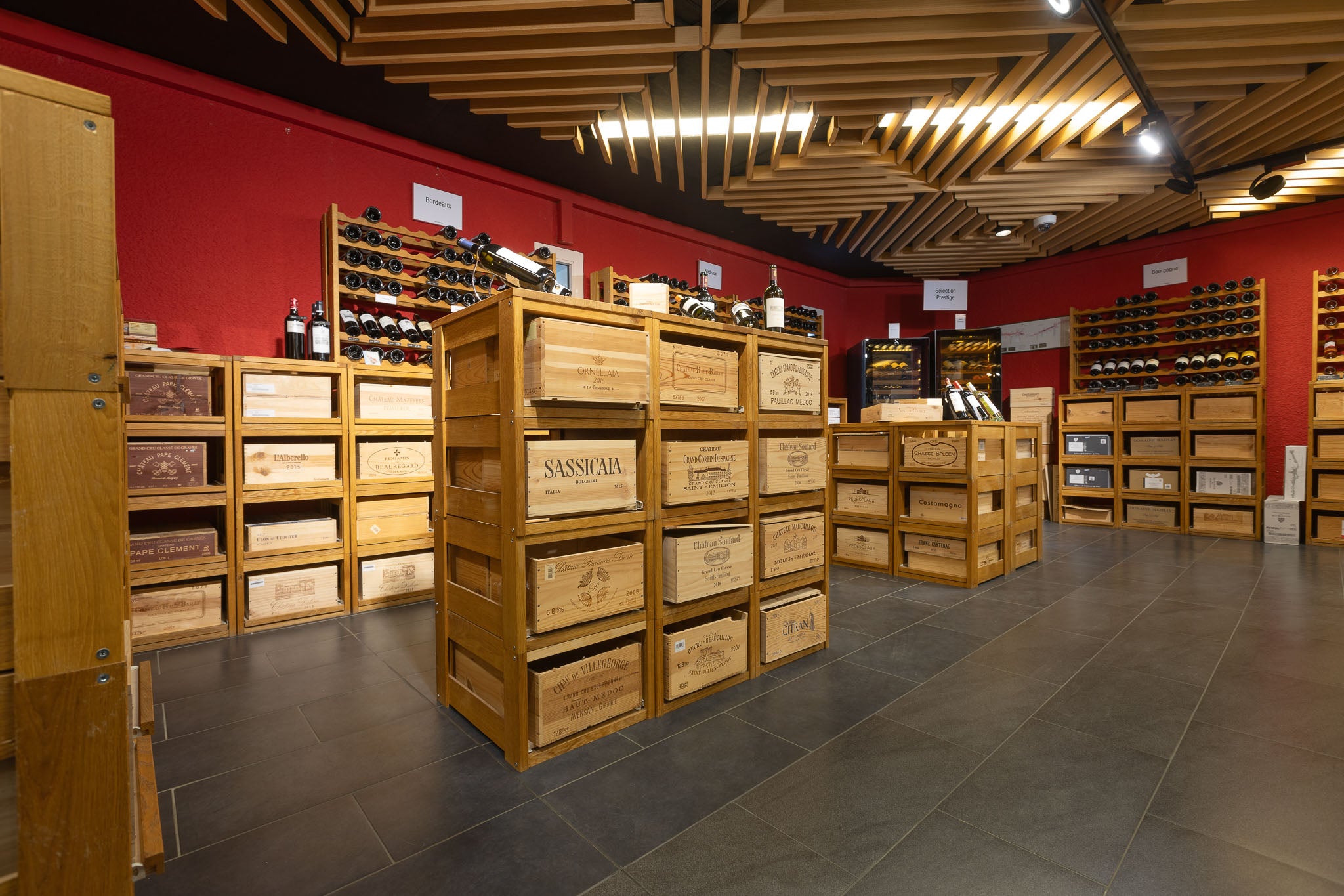 Intérieur d'une vinothèque avec des étagères en bois remplies de caisses en bois estampillées (Sassicaia, Château Souverain, etc.) et de bouteilles, sous un éclairage moderne et des murs rouges.