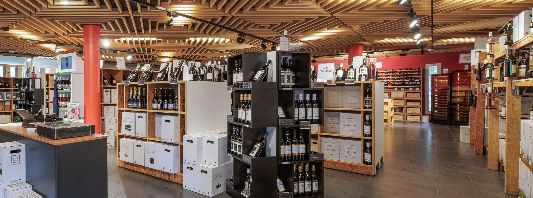Intérieur spacieux et moderne de la vinothèque Cave de La Côte à Tolochenaz, avec un plafond en bois ajouré, des étagères remplies de bouteilles de vin, des caisses en carton empilées et des présentoirs en bois exposant des vins locaux. L'éclairage est chaleureux, mettant en valeur les produits et créant une ambiance accueillante.