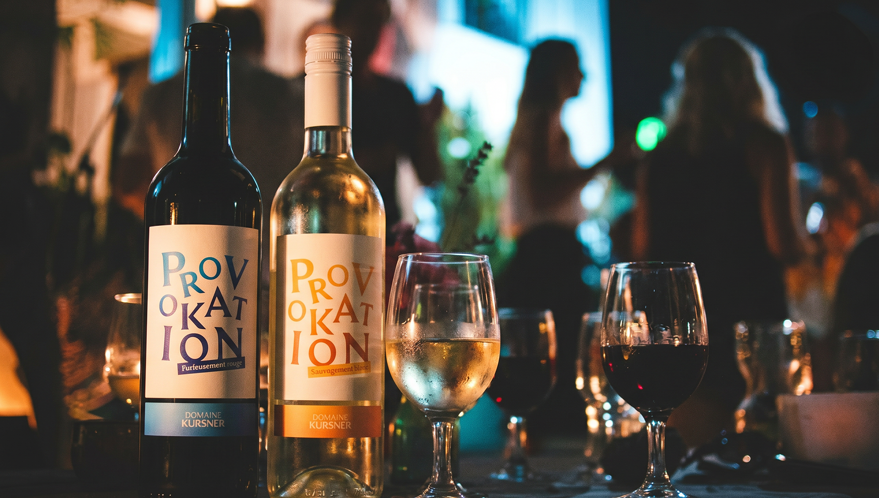 Deux bouteilles de vin du Domaine Kursner étiquetées "PROVOKATION" posées sur une table, accompagnées de deux verres remplis, dans une ambiance de restaurant animé en arrière-plan.