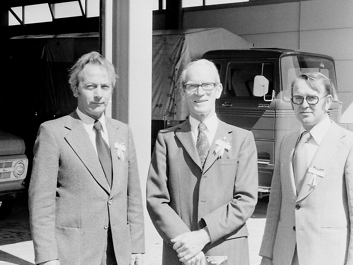 Trois hommes en costumes des années 1970, portant des badges, posant devant un camion devant un entrepôt.