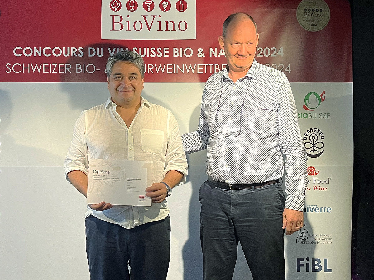 Remise de diplôme du concours BioVino 2024 : Rodrigo Banto, œnologue de Cave de La Côte tient le trophée pour le titre de meilleure Cave Suisse Bio, aux côtés de Julien Hoefliger, CEO, devant une bâche mentionnant le concours du vin suisse bio et nature 2024