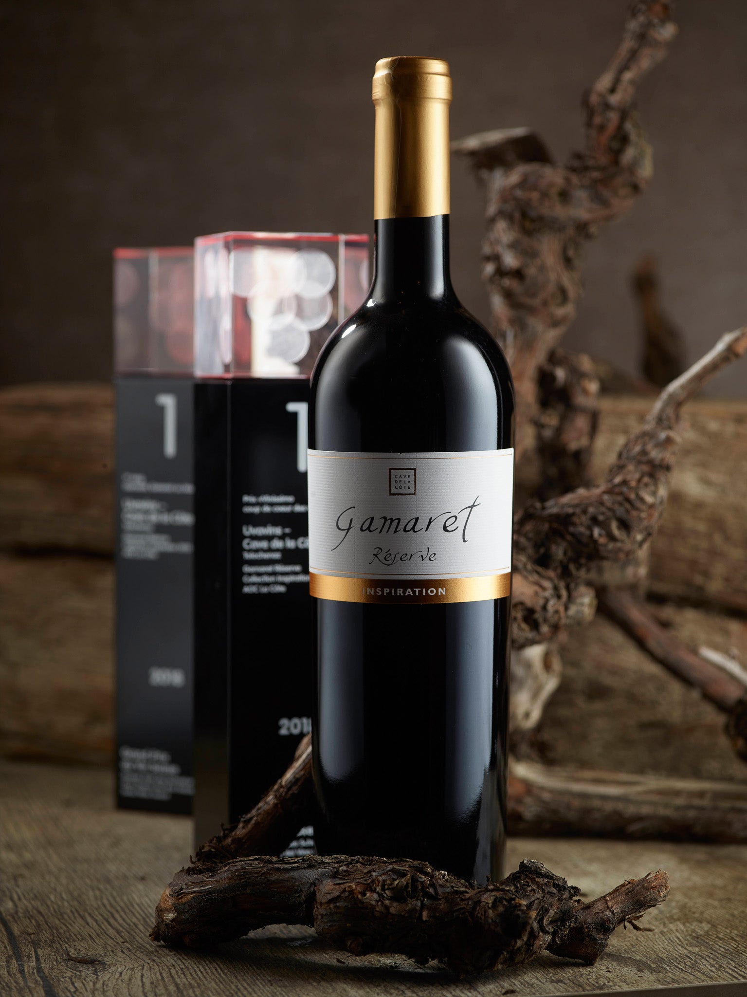 Bouteille de vin rouge 'Gamaret Réserve' de la Cave de la Côte, posée sur une branche de vigne sèche, avec un trophée en arrière-plan.