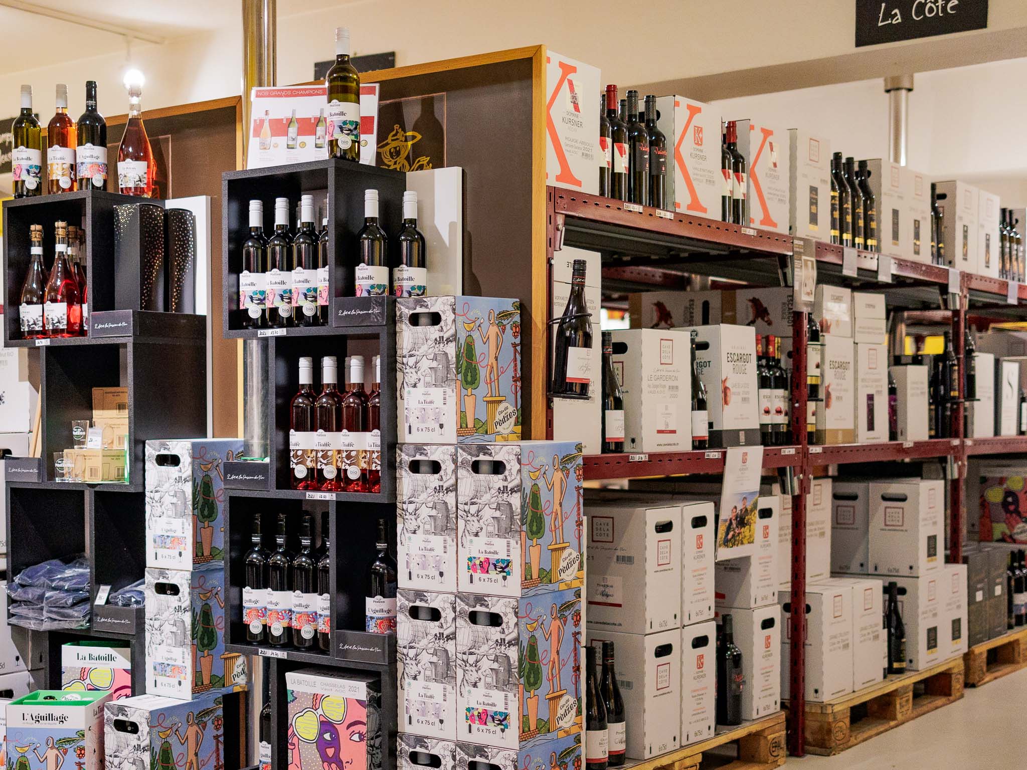 Rayonnages métalliques remplis de bouteilles de vin et de caisses en carton empilées dans la vinothèque Cave de La Côte. Les étagères sont organisées avec des vins de différentes régions, dont des caisses illustrées (ex. : 'La Bâtie', 'L'Aguillage'). L'ambiance est professionnelle et bien rangée, avec une signalétique indiquant 'La Côte' en haut.